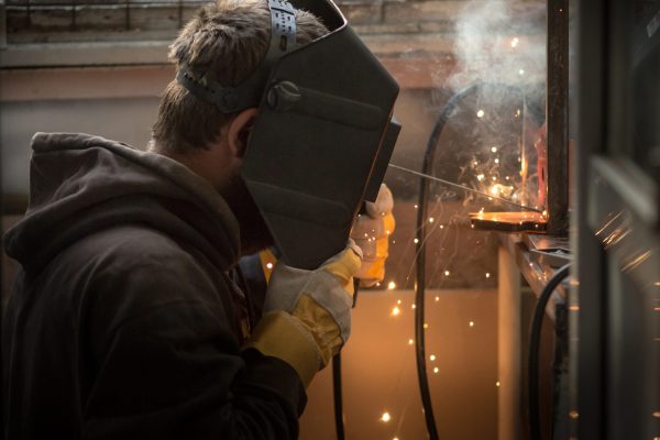 a welder in a welding mask, gloves welds metal with a welding machine and an electrode in his hand, sparks fly, a beautiful glow, bokeh, orange light on a black background, a flash of light and smoke